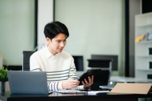 Confident businessman working on finance analysis with a tablet and laptop at office desk.